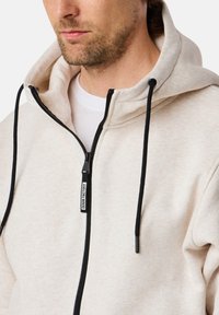 Felpa con zip di un beige chiaro e dalla trama morbida. Presenta lacci neri, una cerniera a contrasto e un semplice etichetta del marchio sul davanti.