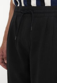 Gros plan d'un pantalon noir à cordon porté avec une chemise rayée bleue, blanche et noire rentrée et boutonnée à la taille.