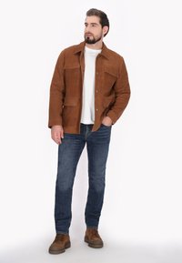 Brun suedejakke med fire lommer, trykknapper og krave, parret med blå jeans og brune snørebåndssko på en hvid baggrund.