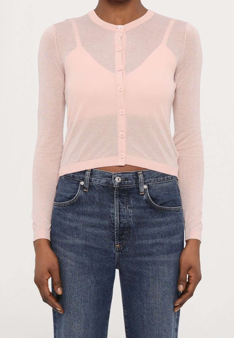 Femme portant un cardigan rose pâle boutonné sur un débardeur assorti et un jean taille haute bleu foncé, debout avec les bras détendus.