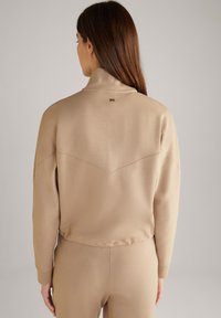 Sudadera deportiva beige de cuello alto con dobladillo ajustado, que presenta un detalle de costura en cheurón y un pequeño logotipo en la parte trasera.