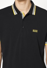 Homme portant un polo noir Hugo Boss avec un col rayé jaune et blanc ainsi que des bordures de manches, trois boutons noirs sur la patte de boutonnage.