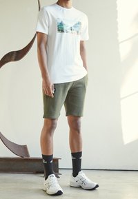 T-shirt bianca in cotone con grafica paesaggistica, abbinata a pantaloni corti verde oliva, sneaker bianche e calze nere con logo.