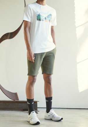 Camiseta blanca de algodón con un gráfico de paisaje, combinada con pantalones cortos verdes oliva, zapatillas blancas y calcetines negros con un logo.