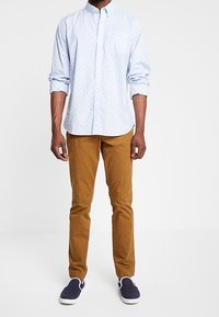 Vaaleansininen nappikauluspaita, jossa on pieniä valkoisia pilkkuja, yhdistettynä ruskeisiin slim fit -housuihin ja tummansiniisiin slip-on-kengiin. Silkkinen puuvillan pinta.
