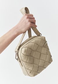 Beige leren handtas met een geweven textuur, ronde vorm, bovenste ritssluiting en een korte handgreep, vastgehouden door een hand tegen een lichte achtergrond.