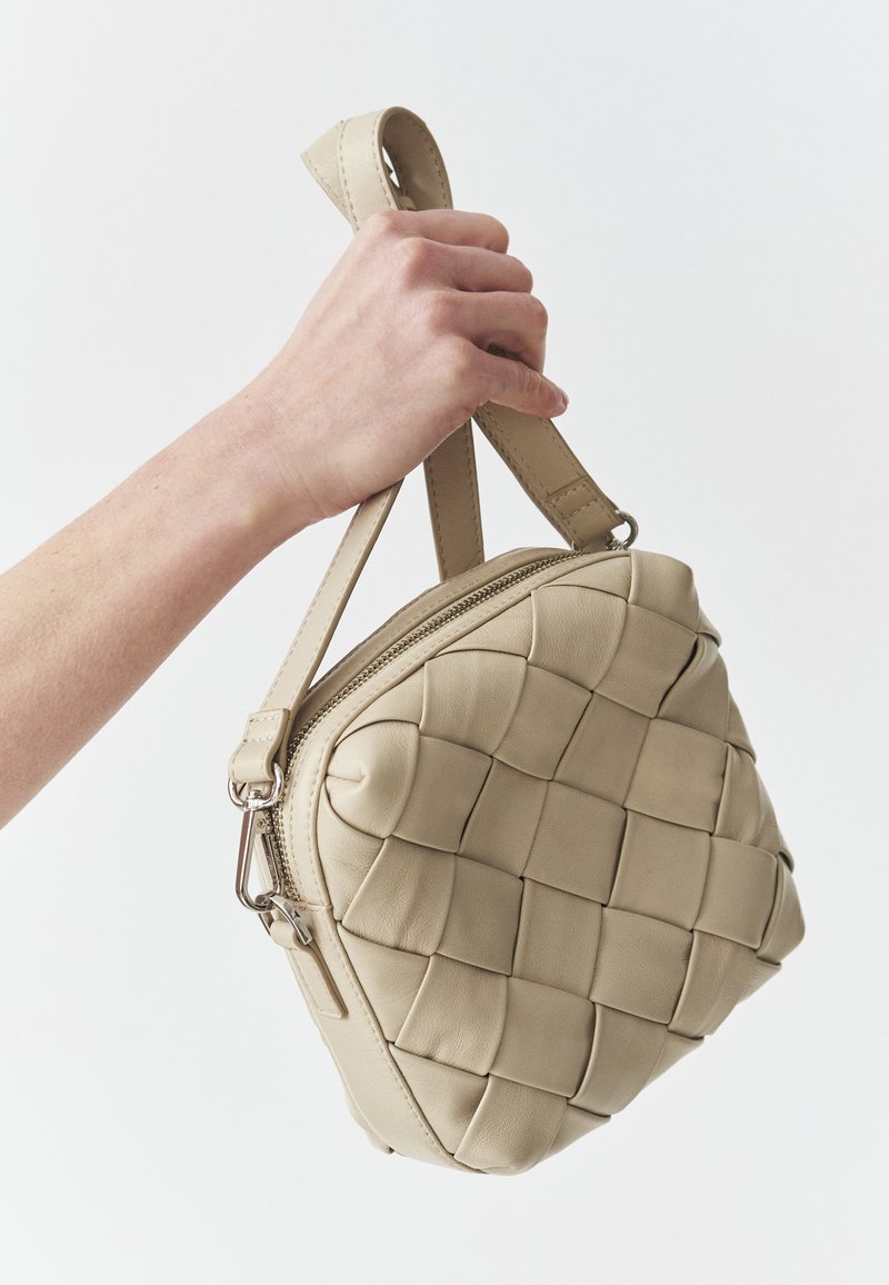 Beige lædertaske med vævet tekstur, rund form, top lynlås og et kort håndtag for nem bæring.