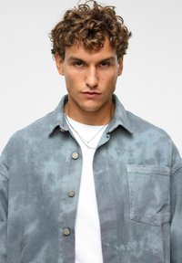 Lichtblauwe denimjack met een vervaagde tie-dye-effect, voorzien van grote knopen, een borstzak en een kraag, over een witte shirt.