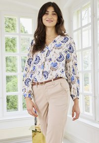 Femme debout à l'intérieur près de grandes fenêtres, portant un chemisier à manches longues à motifs floraux bleus et beiges et un pantalon beige avec une ceinture marron.