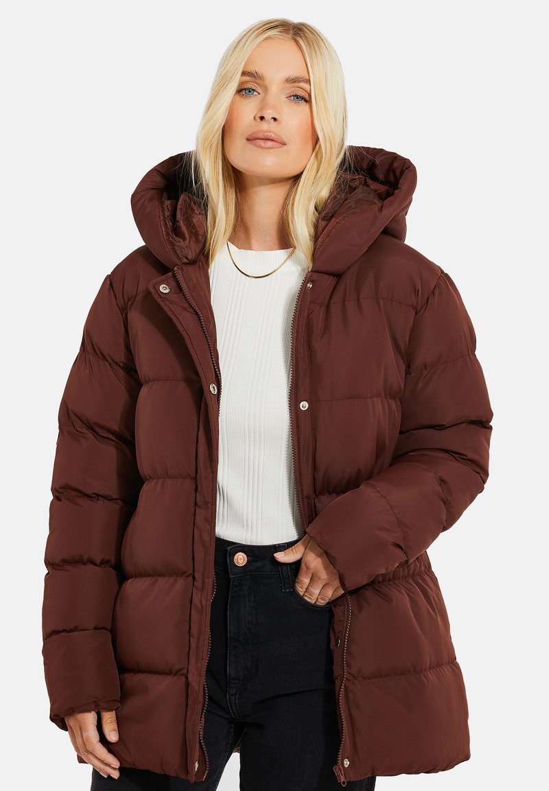 Doudoune marron avec capuche, design matelassé, gros boutons et fermeture éclair. Portée sur un haut à côtes blanc et un jean noir.