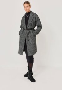 Femme portant un manteau en chevron gris ceinturé sur une robe noire côtelée, des collants transparents, des bottines à lacets noires et un collier en chaîne argenté.