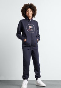 Tuta con zip di colore blu navy realizzata in tessuto morbido, con un logo sul petto, due tasche frontali e polsini elasticizzati. Indossato con sneakers bianche.