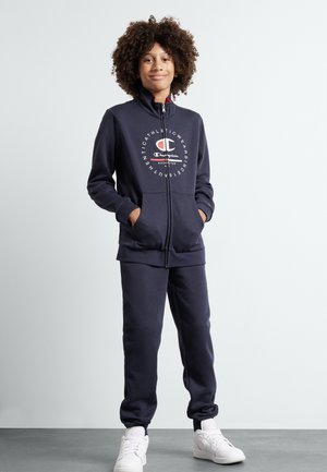 Tuta con zip di colore blu navy realizzata in tessuto morbido, con un logo sul petto, due tasche frontali e polsini elasticizzati. Indossato con sneakers bianche.