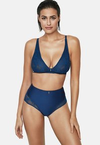 Femme aux cheveux mouillés portant un ensemble coordonné de soutien-gorge en dentelle bleu marine et de culottes taille haute, se tenant contre un fond uni.