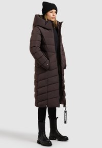 Brauner langer Daunenmantel mit Kapuze und Zickzackmuster; verfügt über Reißverschlusstaschen, einen Reißverschluss und ein schwarzes Etikett. Kombiniert mit schwarzen Stiefeln und Leggings.