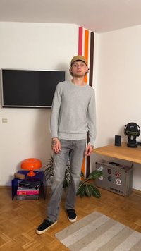 Grau gestrickter Pullover mit gerippten Bündchen, hellblaue Jeans und schwarze Slippers. Der Raum verfügt über bunte Wandstreifen und moderne Dekoration.