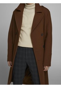 Brauner Wollmantel mit einer doppelt-knöpfbaren Gestaltung, großen Revers und schwarzen Knöpfen. Getragen über einem cremefarbenen Rollkragenpullover und grauen karierten Hosen.