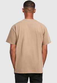 Man with short hair wearing a plain beige short-sleeve T-shirt, viewed from the back against a light gray background.