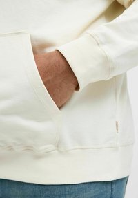 Kremowy sweatshirt z kieszenią kangurzą, ściągaczami na mankietach oraz widoczną metką na dole. Miękka, teksturowana tkanina.