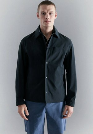 Hombre con chaqueta negra abotonada y pantalones cortos azules, de pie frente a un fondo gris neutro, mirando hacia adelante con expresión neutral.