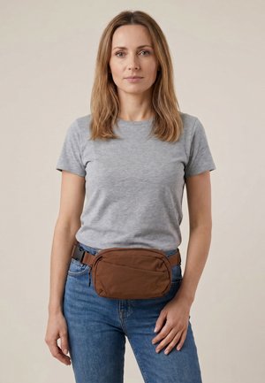 Vrouw met schouderlang lichtbruin haar staat, draagt een grijs T-shirt, blauwe spijkerbroek en een bruine heuptas met een voorvak met ritssluiting.