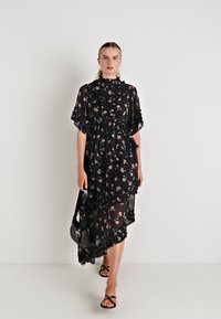 Vestido preto floral com camadas transparentes, bordas onduladas e gola alta. Combinado com uma pequena carteira preta e sandálias com tiras.