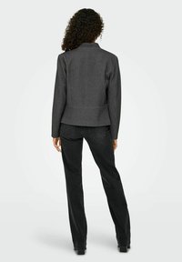 Femme aux cheveux bouclés portant une veste gris foncé et un jean noir à jambes droites, debout de dos sur un fond uni.