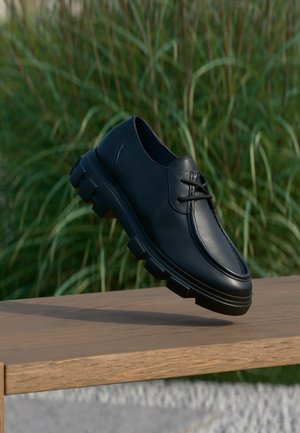 Black leather lace-up shoe with thick tread sole floating above wooden bench with tall green grass background.