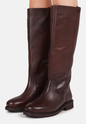 Bottes en cuir marron foncé montant jusqu'aux genoux, avec des talons bas empilés, des bouts ronds et des fermetures éclair latérales, portées sur des jambes nues sur fond blanc.