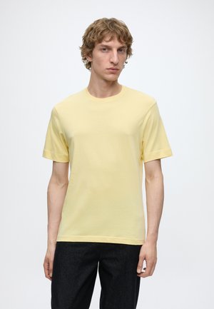 Jeune homme aux cheveux bouclés châtain clair portant un t-shirt jaune uni à manches courtes et un pantalon foncé sur fond blanc.