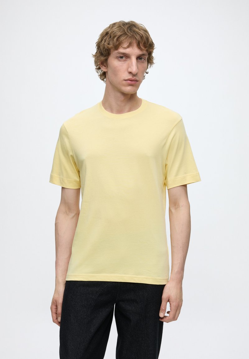 Jeune homme aux cheveux bouclés châtain clair portant un t-shirt jaune uni à manches courtes et un pantalon foncé sur fond blanc.
