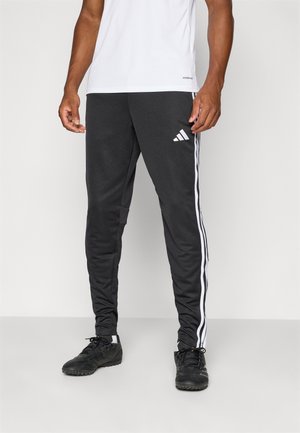 Man draagt zwarte Adidas trainingsbroek met witte strepen en zwarte sportschoenen, staand tegen een effen lichte achtergrond.