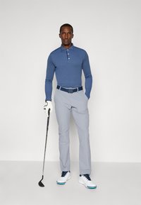 Polo majica z dolgimi rokavi v modri barvi, svetlo sivi hlače, bele čevlje z modrimi poudarki, drži golf palico, nosi belo golf rokavico.