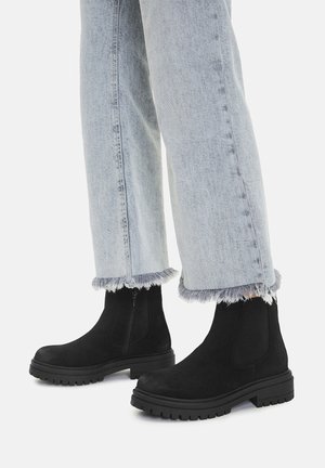 Czarne zamszowe botki na kostkę z masywną gumową podeszwą i zapięciem na zamek, zestawione z jasnoniebieskimi dżinsami o postrzępionych nogawkach.