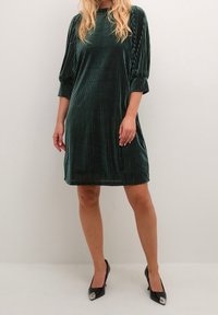 Vestido de terciopelo verde esmeralda con rayas verticales, mangas tres cuartos y corte relajado, combinado con tacones negros con acentos metálicos.