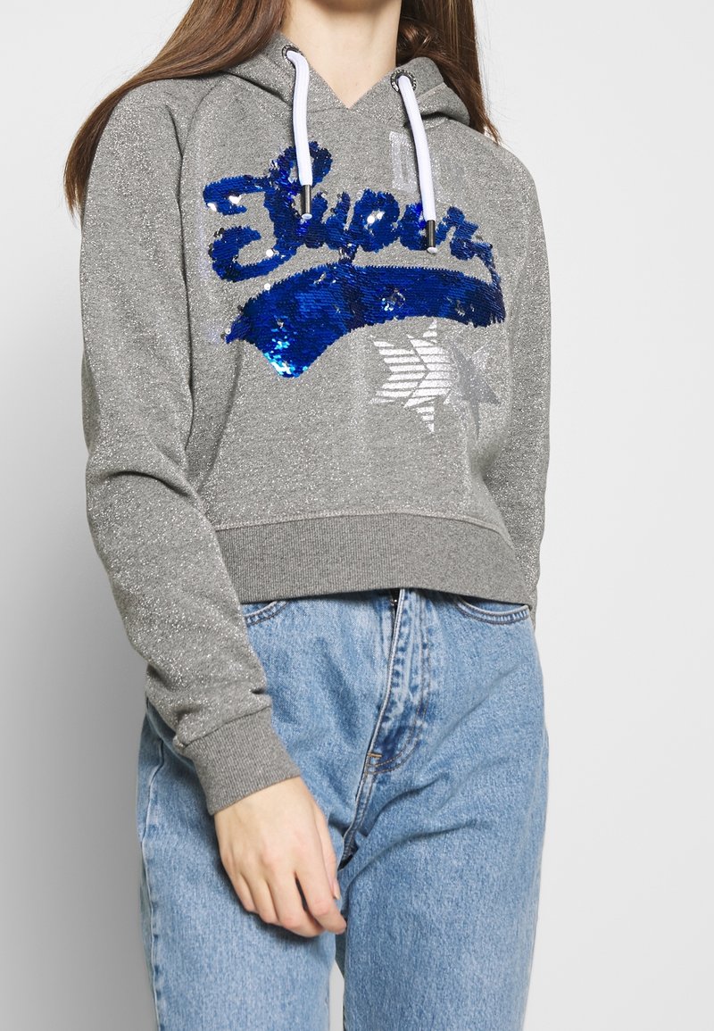 Gray cropped hoodie with sparkly texture, featuring blue sequin lettering and white drawstrings. Paired with light blue denim jeans.