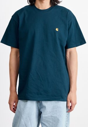 Tumma teelinvihreä puuvillainen t-paita, jossa lyhyet hihat, pyöreä pääntie ja pieni keltainen logo rinnassa. Istuu väljähkösti ja siinä on sileä rakenne.