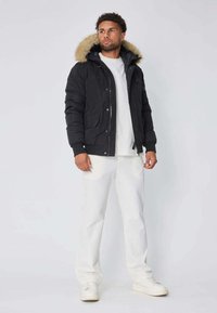 Veste matelassée noire avec une capuche en fourrure, fermeture zippée à l'avant et deux poches avant. Portée sur une chemise blanche et associée à un pantalon blanc et des baskets.