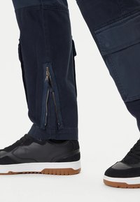 I pantaloni cargo blu presentano gambe inferiori con zip, tasche laterali e una linea dritta, abbinati a scarpe sportive nere con suole bianche.
