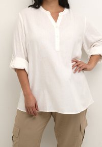 Blouse blanche légère avec un col mandarin et une patte de boutonnage. Présente des manches retroussées. Assortie à un pantalon cargo beige.