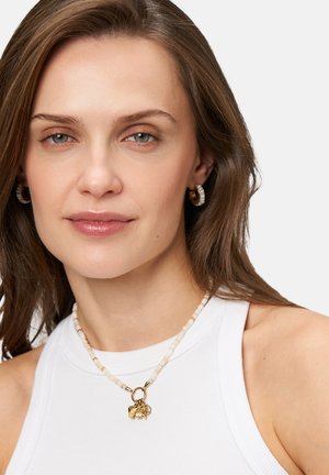 Goldener Charm-Halsreif mit beigen Perlensträngen. Modell trägt ein ärmelloses weißes Top, das die Details der Halskette und die glatte Hauttextur zur Geltung bringt.