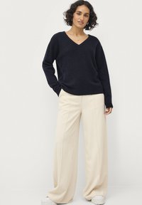 Marineblaues V-Ausschnitt-Pullover aus weichem Strickstoff, kombiniert mit cremefarbenen weit geschnittenen Hosen. Der Pullover hat gerippte Bündchen und Saum, und das Outfit wird durch weiße Sneakers ergänzt.