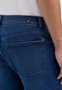 Ciemnoniebieskie dżinsy z denimu o wąskim kroju, posiadające tylne kieszenie oraz małą metkę na prawej kieszeni. Widoczne są również detale przeszyć.