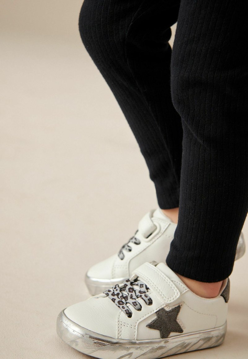 Białe sneakersy z metaliczno-srebrnymi podeszwami, czarnym detalem w kształcie gwiazdy i sznurówkami w leopardzi wzór. Połączone z czarnymi, żebrowanymi legginsami.