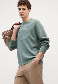 Felpa verde con tessuto testurizzato, dettaglio del logo in rilievo e polsini a coste, abbinata a pantaloni beige e una borsa marrone.