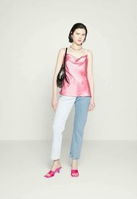 Mulher vestindo uma camisola de cetim rosa, jeans bicolores, sandálias de salto rosa, bolsa preta de ombro, colar prateado, de pé contra um fundo claro e liso.