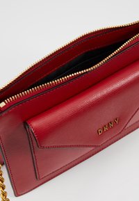 Portafoglio DKNY in pelle rossa con zip dorata e catena, dotato di una tasca frontale in stile busta con lettere logo dorate.
