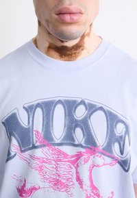 Lys blå bomulds t-shirt med fed grå "NOKE" tekst og et pink vinge-hest grafik over brystet. Blød tekstur, rund hals design.