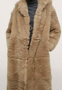 Cappotto in pelliccia sintetica di colore marrone chiaro, con un taglio morbido, cappuccio e tessuto testurizzato con cuciture in vista e senza chiusure.