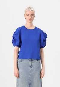 Blue short-sleeve top with textured fabric and puffed sleeves, paired with a denim skirt. The top features a round neckline and casual fit.